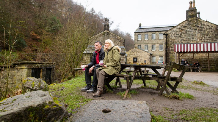 Two visitors sitting on a bench and drinking coffee outside Gibson Mill at Hardcastle Crags, West Yorkshire